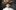 julie fowlis jul 2017 Fowlis wearing a white feather dress and a large brown feather hat, standing in front of a dark gray background.