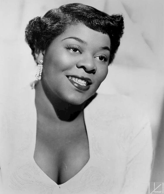 Dinah Washington smiles at camera wearing white top in a black and white image.
